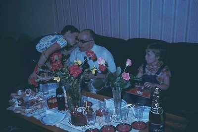 Vintage family portrait from the 1970s featuring three generations at a festive table. Centerpiece includes floral arrangemen...