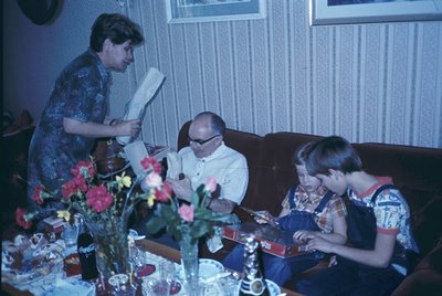 Vintage indoor scene: woman in patterned blouse reads aloud to seated man in white shirt/sunglasses and boy in floral shirt. ...