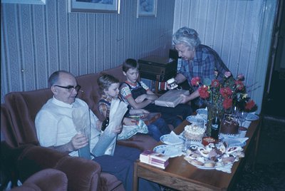 Vintage indoor scene featuring three generations: man in glasses (left), boy in tie-dye shirt (center), and woman in plaid sh...