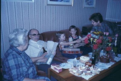 Vintage indoor family gathering, likely 1970s–1980s. Three adults and two children seated around a coffee table laden with a ...