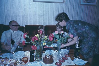 Vintage family celebration with floral centerpieces and cake. Mid-20th century dining room setting featuring a woman assistin...