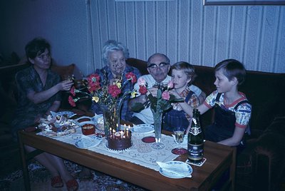 Vintage family gathering around a table with a birthday cake lit by candles, floral centerpiece, and vintage glassware. Mid-2...