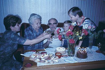 Family celebration with multi-generational toast around a birthday cake (1970s-1980s). Five individuals—adults in patterned b...