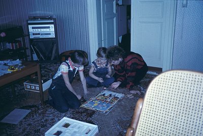 Vintage indoor scene featuring three individuals assembling a pop-culture-themed puzzle on a patterned carpet, likely mid-197...