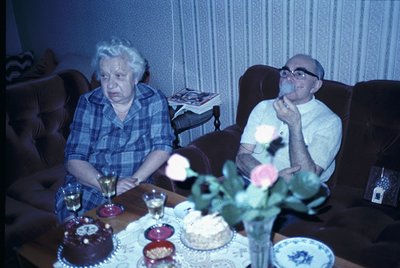 Vintage indoor portrait of an elderly couple seated at a table adorned with two tiered cakes, floral centerpiece, and wine gl...