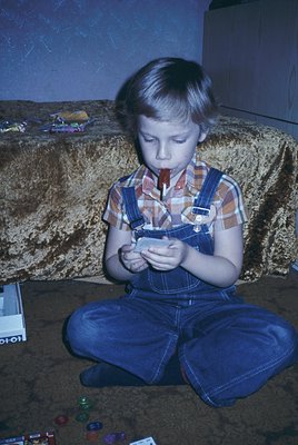 Vintage 1970s-style boy engrossed in a handheld electronic game (Magnavox Odyssey controller) on a worn, patterned couch. Sur...