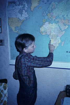 Young boy in plaid shirt pointing at a vintage world map (1960s-70s style) on a classroom wall, likely mid-20th century. Map ...