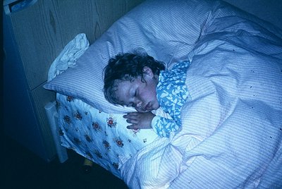 Child sleeping under dim blue light in a simple bedroom, likely 1980s–1990s. Floral-patterned blanket and pillowcase contrast...