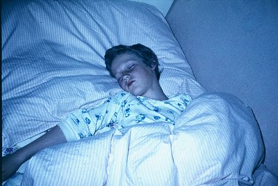 Vintage hospital scene: child resting in bed under dim blue lighting, wearing a floral-patterned hospital gown. Blanket parti...