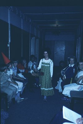 Vintage indoor gathering featuring a woman in a 1970s-style green-and-white patterned dress holding a basket, surrounded by s...