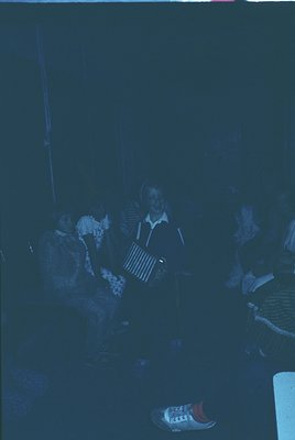 Vintage group photo of a folk music session, likely 1960s–1970s. A man plays an accordion while others sing or clap, seated i...