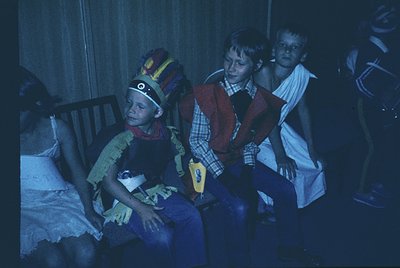 Children in elaborate costumes—one in a colorful, feathered headdress and layered fabric, another in a red vest and plaid shi...