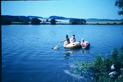 Three individuals paddle a yellow inflatable raft on calm waters, surrounded by lush greenery and distant hills under clear s...