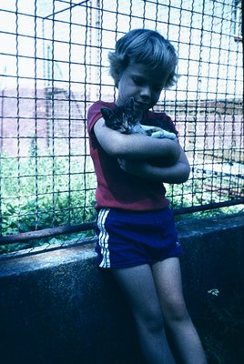 Young boy in 1970s-style red tank top and blue Adidas shorts hugs a striped tabby cat near a chain-link fence. Urban backgrou...