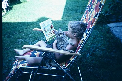 A child in a vintage floral swimsuit reads a children’s book titled *"Puss in Boots"* while a tabby cat rests on their lap in...