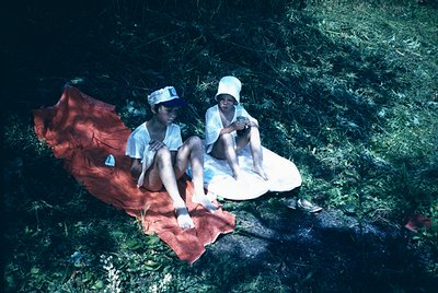 Two children sit on a red blanket near a rocky stream, mid-20th century. Both wear white caps and matching outfits, suggestin...