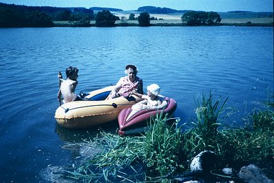 Vintage inflatable raft on calm lake with three adults and a dog, mid-20th century. Bright summer attire suggests leisure act...