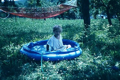 Child in 1970s-style inflatable pool surrounded by lush greenery, with a red woven hammock in background. Mid-century outdoor...