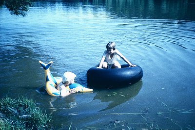 Two children play in shallow water, 1960s-1970s. One sits atop a black inner tube, the other reclines on a yellow inflatable ...