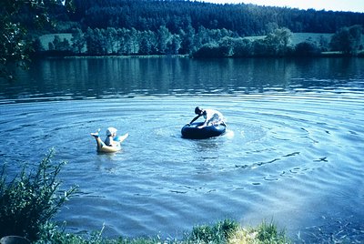 Two individuals float on inner tubes in a serene lake, surrounded by lush greenery and forested hills. The vintage color tone...