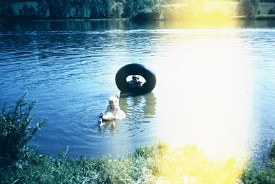 Vintage photo of a person wading in shallow water beside a large, floating tire. Sunlight creates lens flare, suggesting midd...