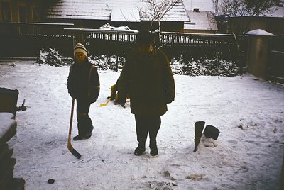Two figures in winter attire clear snow from a residential courtyard, likely mid-20th century. The adult wears a dark coat an...