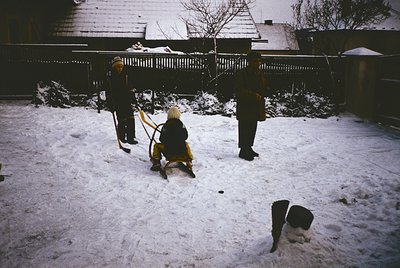 Three children sledding in a residential backyard, mid-20th century. Snow-covered ground, wooden fence, and houses in backgro...