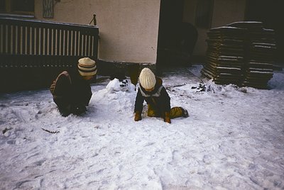 Two children play in deep snow beside a brick building with stacked firewood, likely mid-20th century. Warm winter attire inc...
