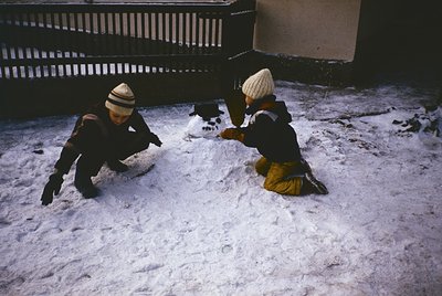Two children play in snow near urban buildings, mid-20th century. Both wear winter gear: hats, gloves, and heavy coats. One k...