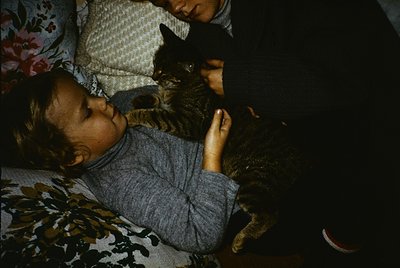 Vintage sepia-toned photo of two children and a cat sharing a bed. Child on left wears a striped shirt and headscarf, resting...