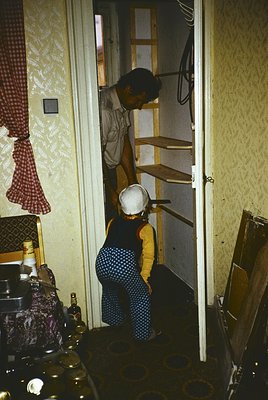 Mid-20th century kitchen scene featuring two women—one standing on a ladder holding a metal pot, the other holding a child in...