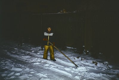 Child in winter attire clearing snow beside a fence, likely in a residential area. Yellow rubber boots and heavy jacket sugge...