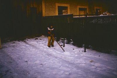 A lone figure in winter attire shovels snow in a dimly lit alleyway, framed by a yellowish building with barred windows. The ...