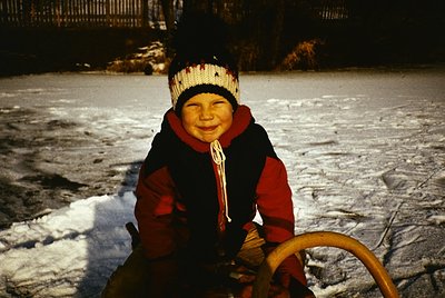 Vintage winter scene of a child in a vintage-style knit hat and red/black winter coat, seated on a wooden sled. Snow-covered ...