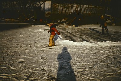 Street scene at dusk: two workers in winter gear clear snow with shovels and a push broom. Urban setting with industrial ligh...