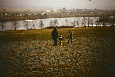 Vintage sepia-toned photo of two adults and a child walking across a frosted field, leading a dog. Distant village and lake r...