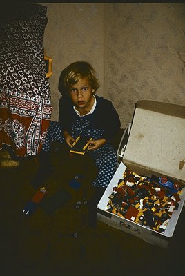 Mid-20th-century child playing with Lego-style bricks in a dimly lit room, likely 1950s–1960s. Open box reveals classic molde...