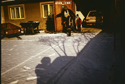 Vintage winter scene featuring a residential courtyard with snow-covered ground. Two figures in winter clothing—one shoveling...