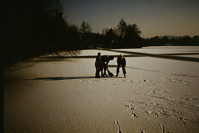 Three figures in winter gear stand on a frozen lake, casting shadows on cracked ice. Low-angle sunlight illuminates their sil...