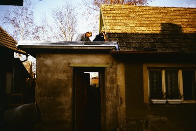 Two individuals repair a rustic clay-tiled roof on a weathered adobe house. The scene captures rural manual labor, likely in ...