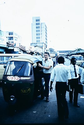Vintage urban street scene featuring a man in a light-colored shirt exiting a yellow SEP auto-rickshaw () in a bustling city....