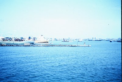 Vintage coastal port scene with industrial docks, cranes, and ships. Urban skyline in background suggests a mid-20th-century ...