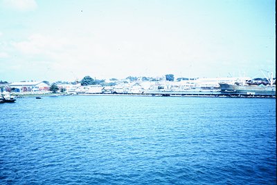 Vintage coastal port scene with low-lying buildings and industrial structures along a waterfront. Prominent pier extends into...