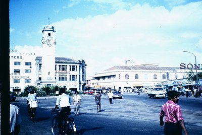 Vintage urban scene featuring a prominent clock tower with "Bulters Cycles" signage on a multi-story building, likely a 1960s...