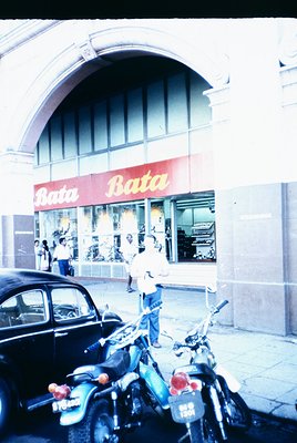 Vintage 1960s-era Bata shoe store with arched entrance, featuring bold red signage and large display windows. Classic cars an...