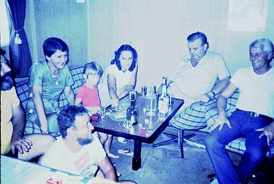 Vintage family gathering in a casual indoor setting, likely mid-1970s. Six adults and one child seated around a wooden table ...