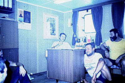Vintage indoor scene featuring a seated speaker at a wooden podium in a modestly furnished room, likely a 1970s-era office or...