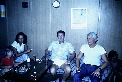 Vintage indoor family portrait in a retro diner-style setting, 1970s. Three adults and a child seated around a round table wi...