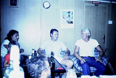 Vintage group photo in a retro hotel room, likely 1970s–1980s. Three adults seated on a bed, smiling at the camera, with a wo...