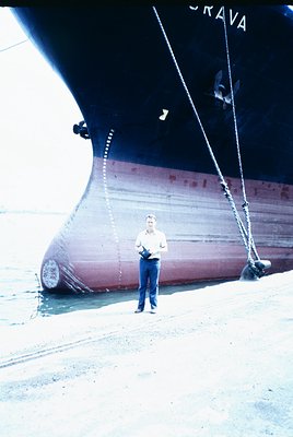 Mid-20th century cargo ship ("GRAVA") docked at port, with a man in light-colored shirt and dark pants standing beside its hu...